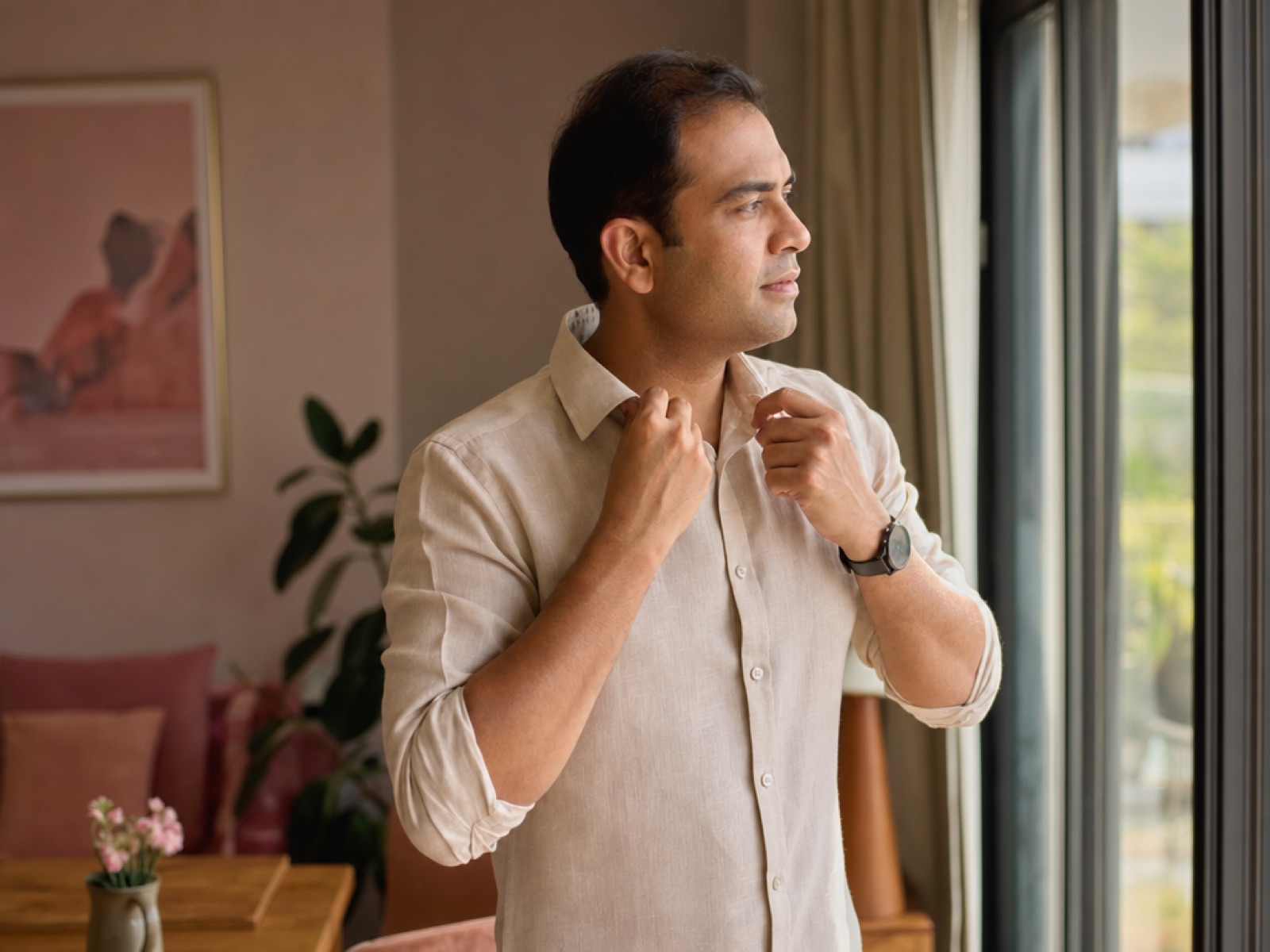Indian man in a linen shirt adjusting his collar by a sunlit window at home